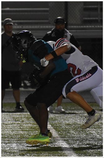 Two people in football uniforms are engaged in a tackle on a grassy field under stadium lights. Description generated by AI