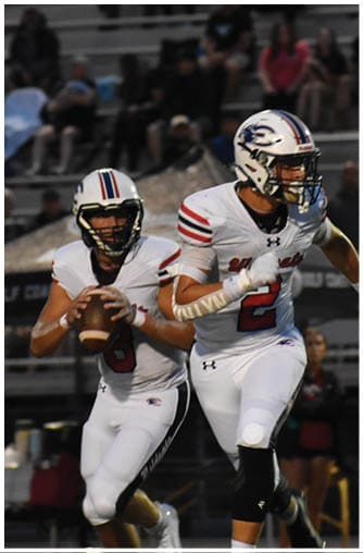 Two people in white football uniforms with helmets are on a field, one holding a football ready to pass while the other runs ahead. Description generated by AI