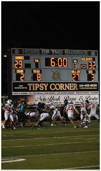 A scoreboard shows a tied football game with six minutes remaining while players in helmets and uniforms engage in a play on the field under stadium lights. Description generated by AI