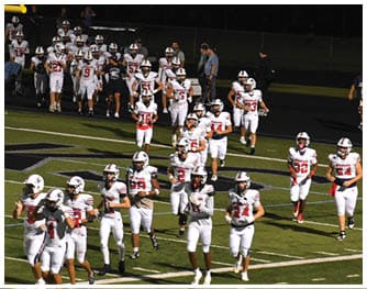 A group of people wearing white football uniforms with helmets are walking on a football field under stadium lights. Description generated by AI