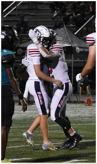 Two people wearing white football uniforms with black and red accents are standing on a grassy field during a game. Description generated by AI