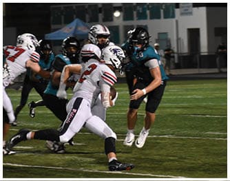 A group of people wearing football uniforms are actively playing on a field at night with one person running while holding the ball and others attempting to block or tackle. Description generated by AI