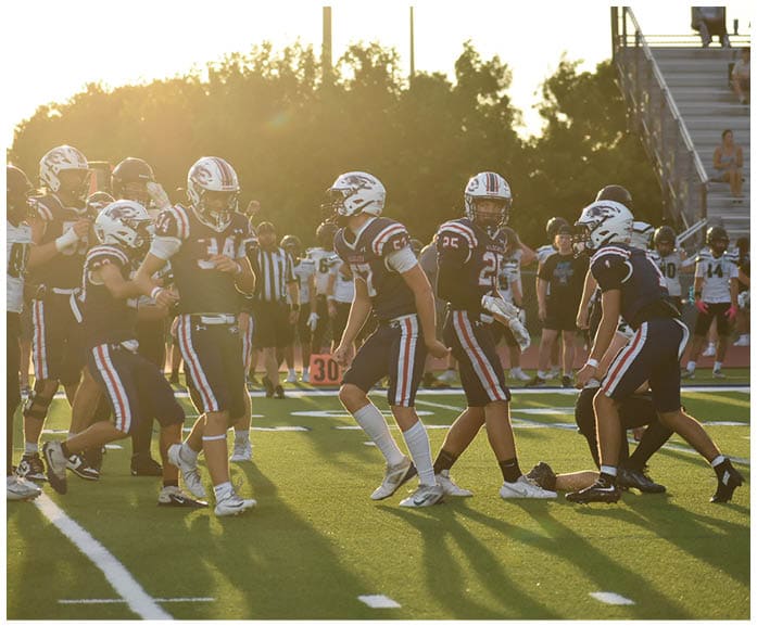 People wearing sports uniforms and cleats are walking and standing on a sunlit grassy field with white yard lines. Description generated by AI