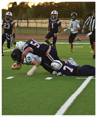 Two people in football uniforms are on the ground during a game, with one wearing a white helmet and the other a dark helmet. Description generated by AI