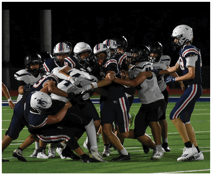 A group of people wearing football uniforms are engaged in a close physical play on a well-lit football field at night. Description generated by AI