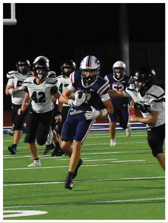 A group of people wearing football uniforms are playing on a well-lit field at night with one person running with the ball while others attempt to block or tackle. Description generated by AI