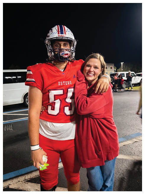 A person wearing a red football uniform and helmet stands next to another person in a red sweatshirt who is smiling and leaning on their shoulder against a dark background. Description generated by AI
