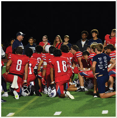 Players in football uniforms are lined up on the field preparing for a play with spectators seated sparsely in the bleachers behind them at night. Description generated by AI