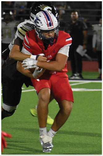 Two people in football uniforms are engaged in a tackle on a grassy field under stadium lights. Description generated by AI