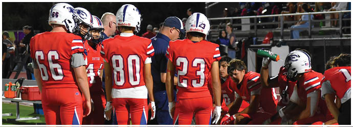 A group of people in white football uniforms with helmets are lined up on a field preparing for a play against opponents in dark uniforms during a nighttime game. Description generated by AI