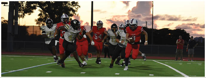 People in football uniforms are actively engaged in a game on a field with one person running with the ball while others block and tackle. Description generated by AI