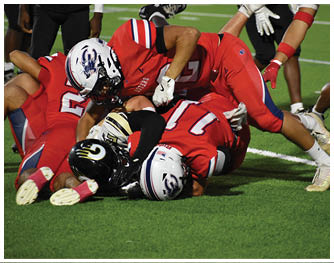 A group of people wearing football uniforms are actively playing on a field at night with one person running while holding the ball and others attempting to block or tackle. Description generated by AI