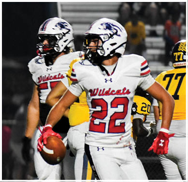 Two people in white Wildcats football uniforms are playing against two people in yellow and black football uniforms on a field with bleachers in the background. Description generated by AI