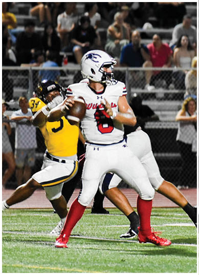 A person in a white football uniform with red socks is preparing to throw a football while being closely defended by a person in a yellow and black uniform on a football field with spectators in the background. Description generated by AI