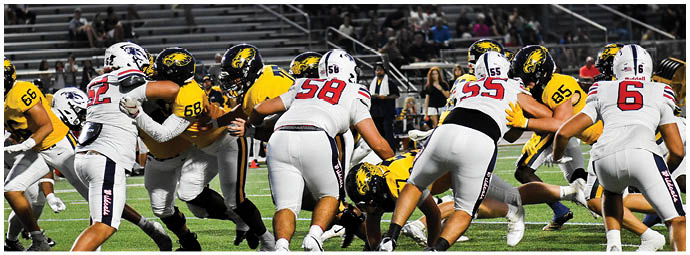 Two opposing American football teams in yellow and white uniforms are lined up facing each other on the field during a game with spectators in the background. Description generated by AI