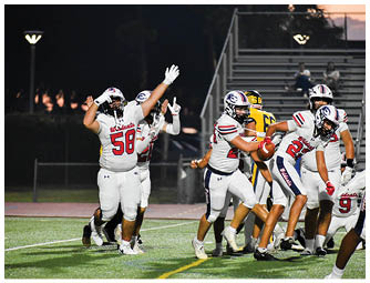 Several people in football uniforms are actively playing on a field at night with one person holding the ball and others raising their arms. Description generated by AI