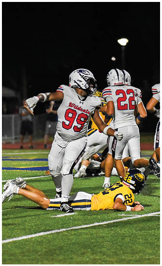A football player in a white uniform with the number 99 is standing over a player in a yellow uniform lying on the field during a nighttime game. Description generated by AI