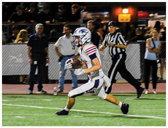 A person in a white football uniform is running on a field holding a football while other players and spectators watch in the background. Description generated by AI