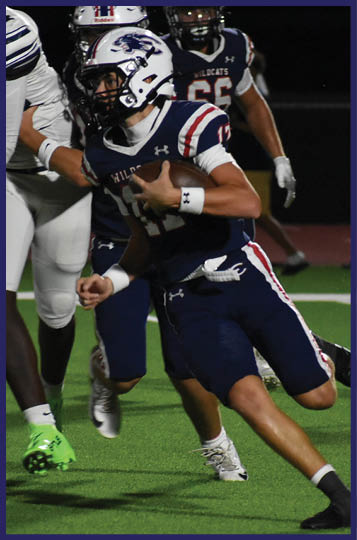 A person in a dark blue football uniform is running with the ball while being pursued by players in white and dark uniforms on a green field at night. Description generated by AI