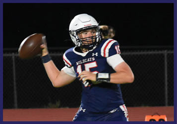 A person in a football uniform is preparing to throw a football on a field while other players and coaches stand nearby during a nighttime game. Description generated by AI