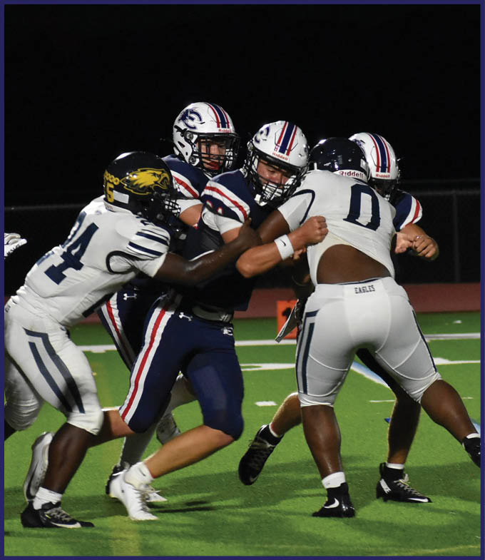 Two football players in dark uniforms are being tackled by two players in white uniforms on a green field during a nighttime game. Description generated by AI