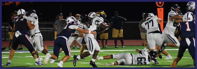 People in football uniforms are actively playing on a field at night with one player running while others block or tackle. Description generated by AI