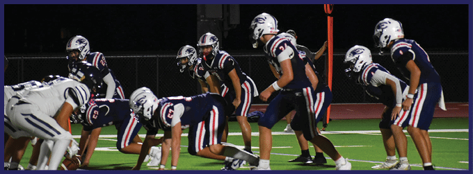 Two opposing American football teams are lined up facing each other on a field, preparing for a play during a game. Description generated by AI