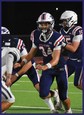 People in football uniforms are actively engaged in a game on a green field under stadium lights at night. Description generated by AI