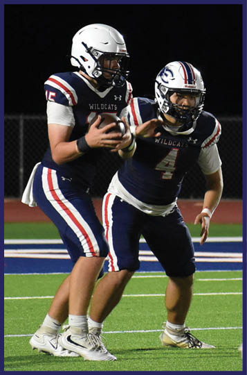 Four football players in helmets and uniforms are on a field at night with one player holding the ball preparing to pass while others are positioned for a play. Description generated by AI