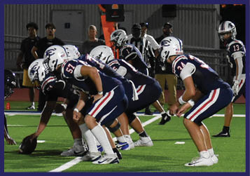 Players in football uniforms are lined up on a field at sunset preparing for a play. Description generated by AI