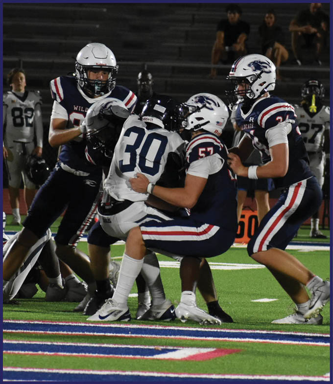 A group of football players in dark and white uniforms are engaged in a tackle on a brightly lit field with spectators in the background. Description generated by AI
