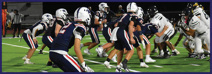 Two opposing American football teams in uniforms are lined up on a well-lit field at night preparing for a play. Description generated by AI