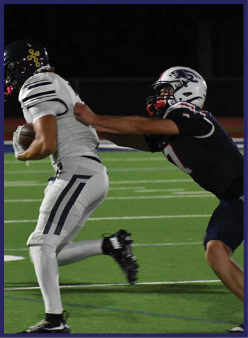 Two people in football uniforms are on a field at night with one person running while holding the ball and the other reaching to tackle them. Description generated by AI
