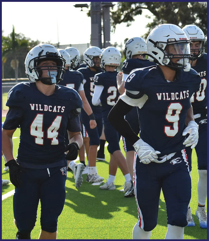 A group of people wearing Wildcats football uniforms and helmets are standing on a field with one person in a referee uniform gesturing in the background. Description generated by AI