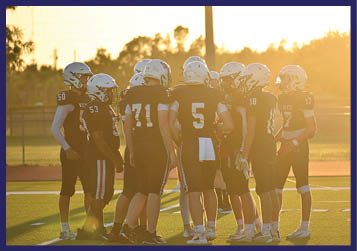 Players in football uniforms are lined up on a field at sunset preparing for a play. Description generated by AI