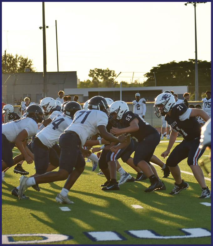 A group of football players in dark and white uniforms are engaged in a tackle on a brightly lit field with spectators in the background. Description generated by AI