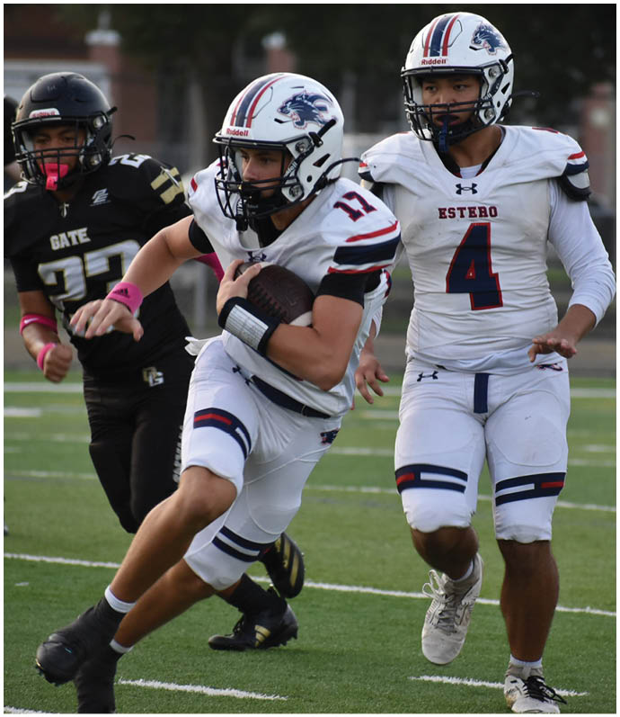 Two people in white football uniforms with helmets are running on a field while being pursued by players in black uniforms. Description generated by AI