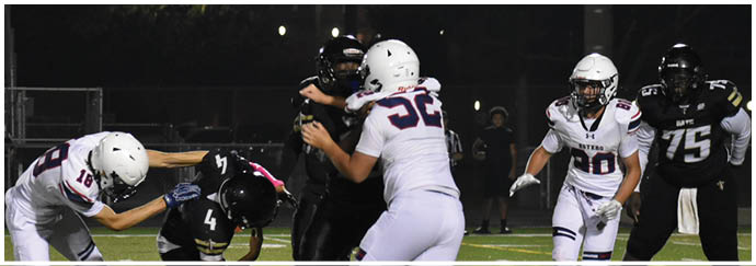 People wearing football helmets and uniforms are engaged in a tackle on a lit outdoor football field at night. Description generated by AI
