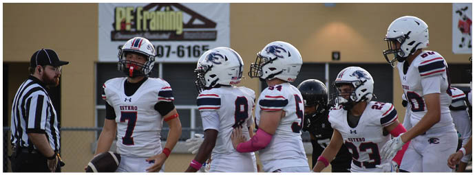 A group of football players in white uniforms with helmets are gathered near a referee wearing a black and white striped shirt on a sports field. Description generated by AI