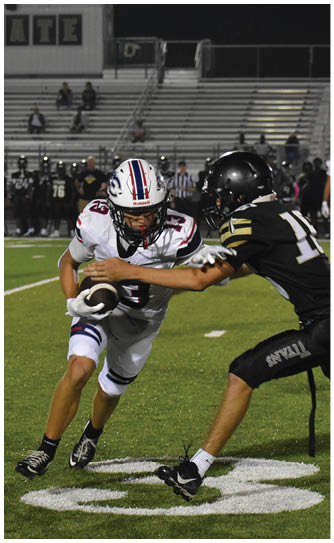 An adult football player in a white uniform is running with the ball on a green field while a referee and other players stand in the background near empty bleachers. Description generated by AI