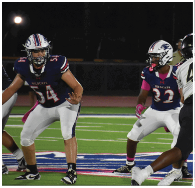 Two people in football uniforms are engaged in a play on a field, with one wearing a white jersey numbered 4 and the other in a dark jersey, while a third person in a blue and white uniform with the word Wildcats watches nearby. Description generated by AI