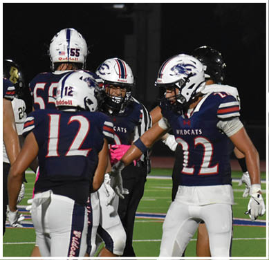 People wearing football uniforms are gathered on a field at night, with some players interacting and one standing apart facing away. Description generated by AI