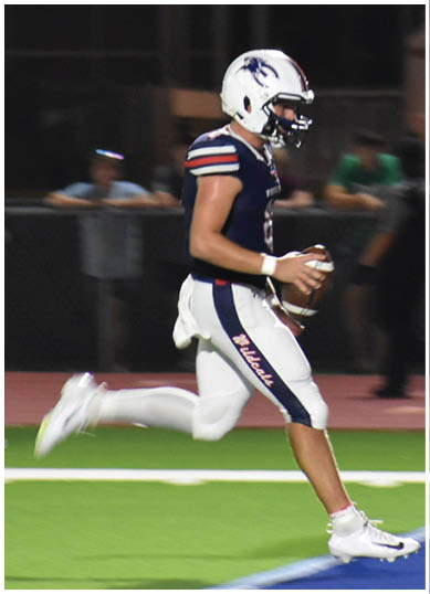 A person in a football uniform is holding a football and preparing to throw it while another person in a different uniform raises an arm on a sports field at night. Description generated by AI