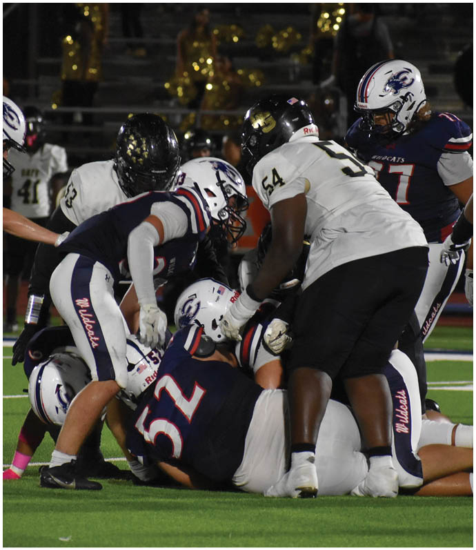Several people in football uniforms are engaged in a play on a green field with some players on the ground and others standing or crouching around them. Description generated by AI