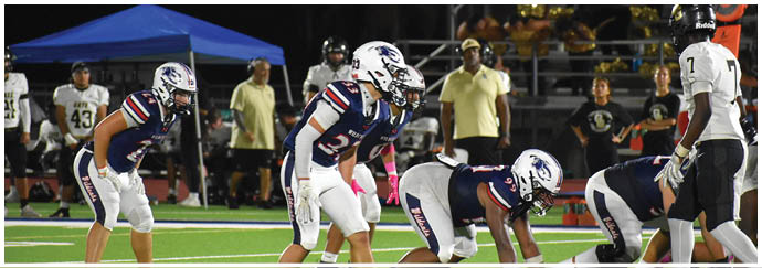 Several people in football uniforms are positioned on a field facing an opposing player in white, with spectators and a blue canopy visible in the background. Description generated by AI