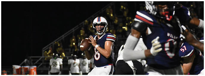 A person in a dark football uniform prepares to throw a football while other players and a marching band are visible in the background on a nighttime field. Description generated by AI