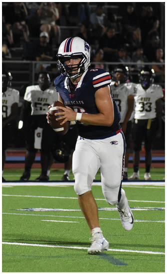 A person wearing a football uniform with a helmet is running on a field while holding a football, with other players and spectators visible in the background. Description generated by AI