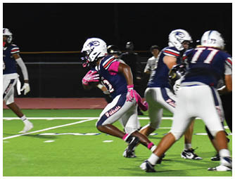 A group of people wearing football uniforms are playing on a well-lit field at night with one person running while holding the ball. Description generated by AI
