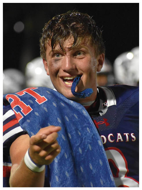 A person with wet hair and black face paint under their eyes is wearing a white sports jersey and appears focused or tired against a dark background. Description generated by AI