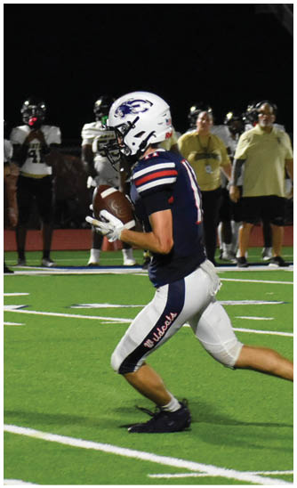 A person in a football uniform is running on a field at night while holding a football, with other people and a referee visible in the background. Description generated by AI